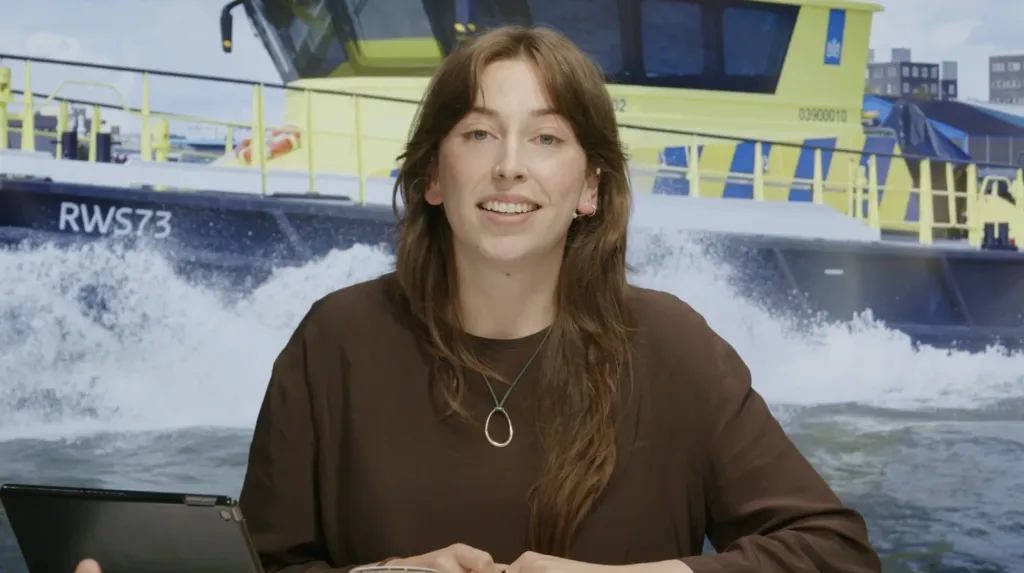 Presentator in de webinarstudio, met een Rijkswaterstaat-vaartuig op de achtergrond