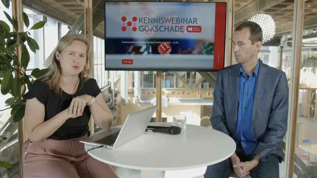 Twee personen bij de statafel met de openingsslide Kenniswebinar Gokschade op het scherm