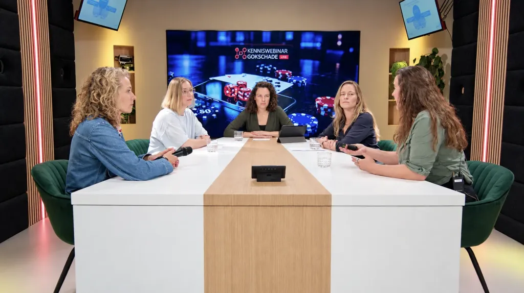 Vijf sprekers aan de tafel in de webinarstudio tijdens het Kenniswebinar Gokschade, met het webinarbeeld op het grote scherm erachter